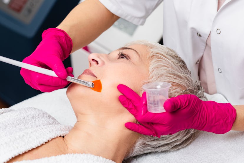 Beautiful senior woman having chemical peeling beauty treatment. The expert beautician is applying c...
