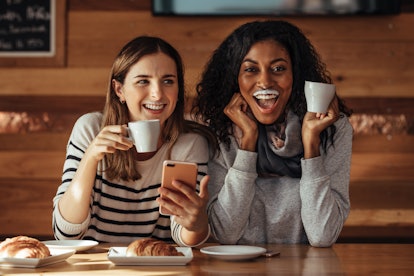 Two women drinking coffee and smiling with a milk mustache in coffee shop. Friends sitting at a cafe...