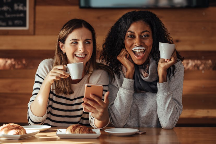 Two women drinking coffee and smiling with a milk mustache in coffee shop. Friends sitting at a cafe...