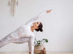 A woman dressed in workout pants and a long-sleeve top strikes a yoga pose at home in a bright room.