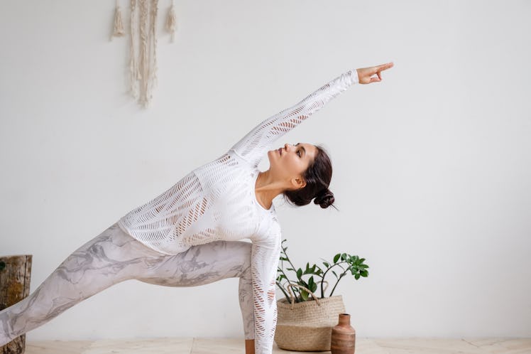 A woman dressed in workout pants and a long-sleeve top strikes a yoga pose at home in a bright room.