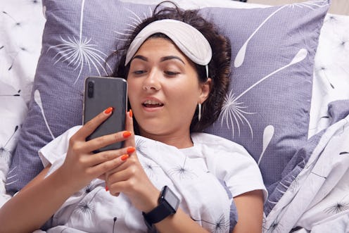 Indoor shot of happy young woman texting on mobile phone or having video call while lying in bed und...