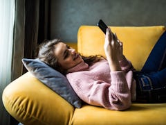 A woman in a pink sweater laying down on her yellow couch, looking at her phone. 