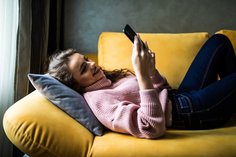 A woman in a pink sweater laying down on her yellow couch, looking at her phone. 