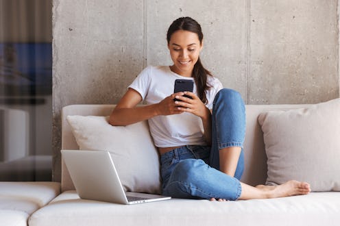 A happy woman sits on her couch with her laptop computer, while looking at her phone.