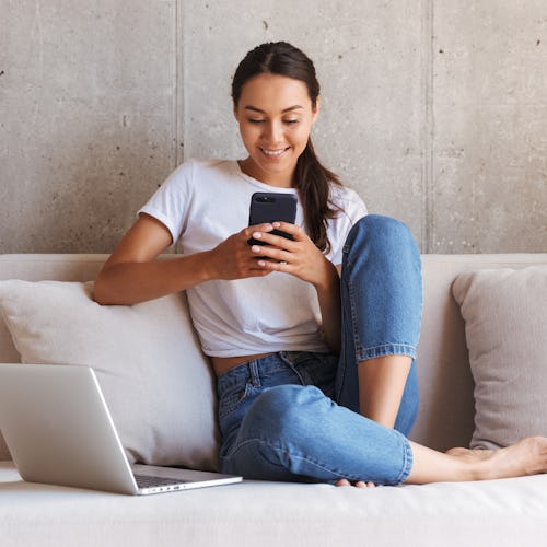 A happy woman sits on her couch with her laptop computer, while looking at her phone. 