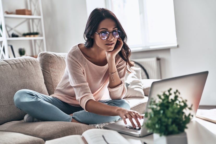 A woman wearing glasses, jeans, and a pink sweatshirt types on her computer while sitting on a gray ...
