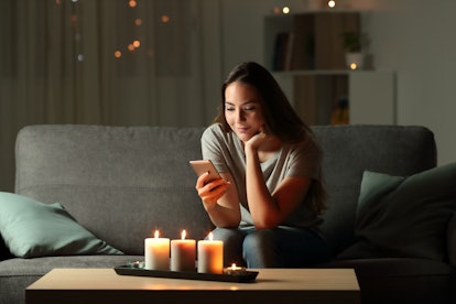 Relaxed girl using phone in the night with candle lights sitting on a couch in the living room at ho...