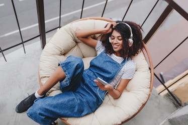 A woman wearing headphones smiles and relaxes on a chair while holding her phone.