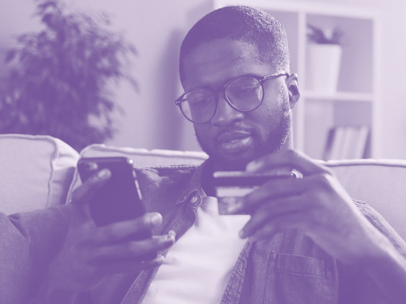 African young man in glasses shopping online with credit card using smart phone at home. Indoor.