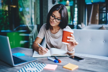 Concentrated female student writing in notebook while learning with cardboard coffee cup in cafe, pe...