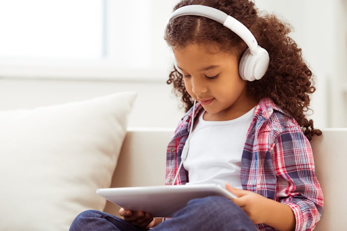 Cute little Afro-American girl in casual clothes and headphones using a tablet and listening to musi...