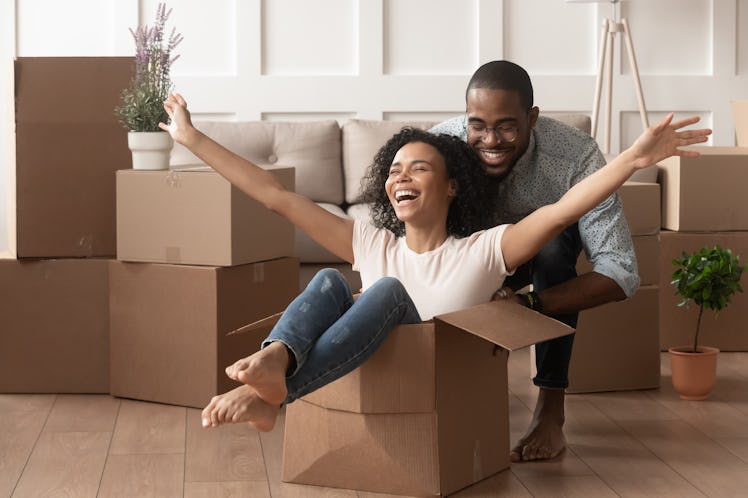 Two young Black people laugh in their new home, surrounded by cardboard boxes. Moving during COVID c...