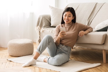 Happy Asian Girl Messaging on Phone at Home, Sitting on Floor near Sofa