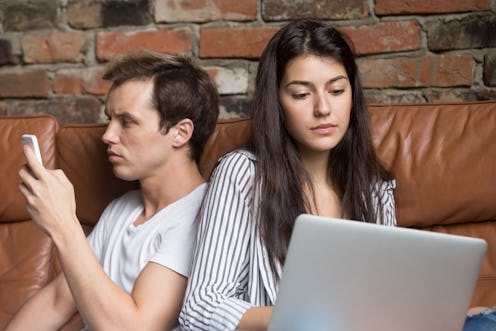 Young couple sitting back to back at home on couch addicted obsessed with modern gadgets, hipster gu...