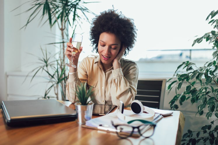 A young woman sits at her kitchen table with a laptop, phone, and glass of wine.