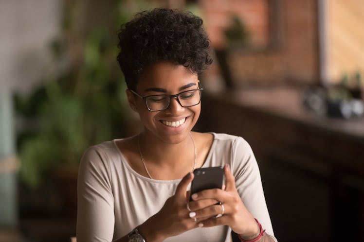 Happy millennial african american woman holding smartphone using ecommerce apps, playing mobile game...