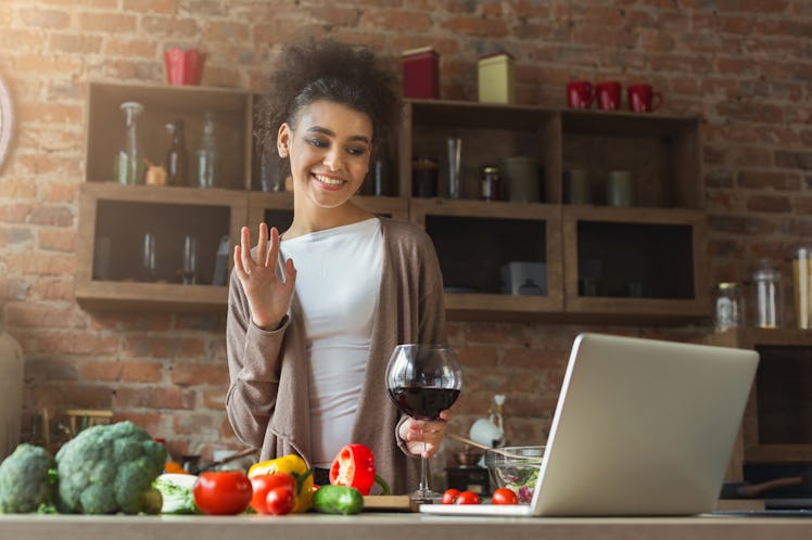 A happy woman standing in her kitchen at home waves at her computer and holds a glass of wine during...