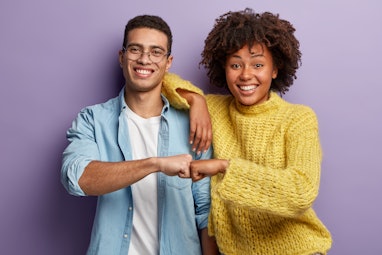 Young diverse couple give fist bump, agree to bring plan to life, smile broadly, have mixed race rel...