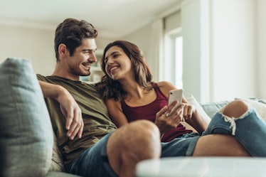 A couple sitting on the couch, smile at each other while looking at a cell phone. 