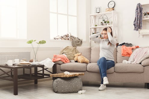 Desperate helpless woman sitting on sofa in messy living room. Young girl surrounded by many stack o...