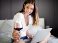A brunette woman smiles and holds a glass of red wine and laptop on her lap.