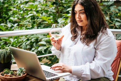 A woman sitting on her balcony outside, enjoys a glass of wine during a virtual wine tasting on her ...