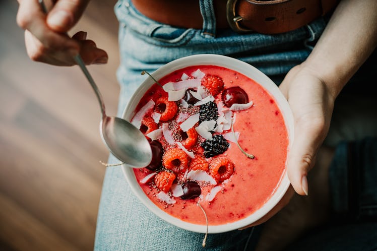A young woman sits with a bright red smoothie bowl in her lap with fresh fruit on top.