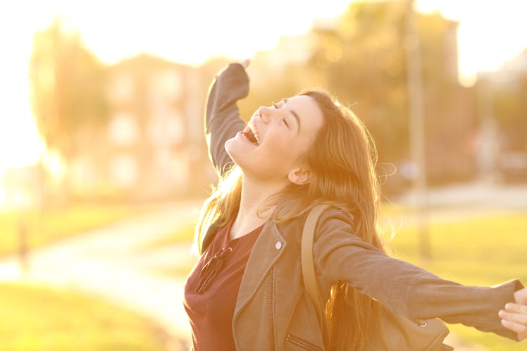 Portrait of an excited teenager girl raising arms and laughing in the street at sunset with a warm l...