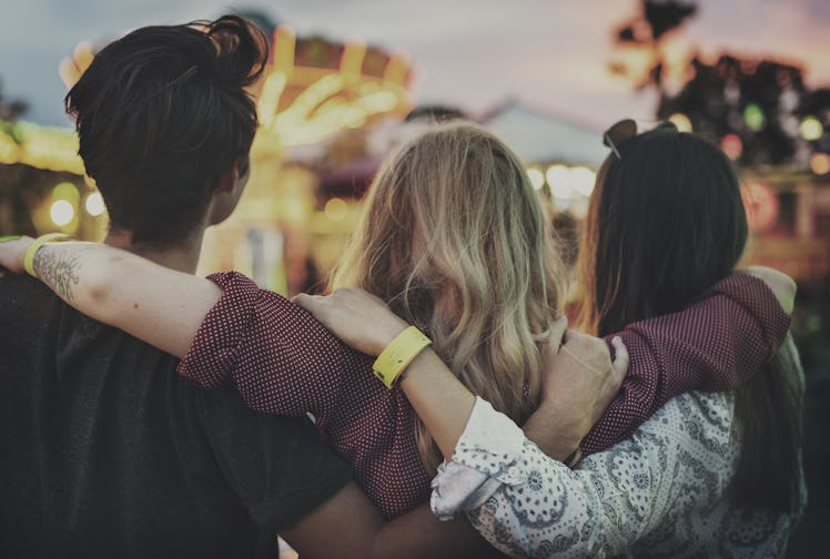 Three friends put their arms around each other and look at a ride at a theme park at dusk.