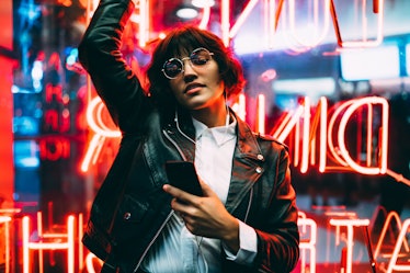 A woman in a leather jacket and sunglasses, dances in front of a neon sign while holding her phone d...