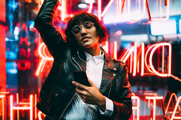 A woman in a leather jacket and sunglasses, dances in front of a neon sign while holding her phone d...