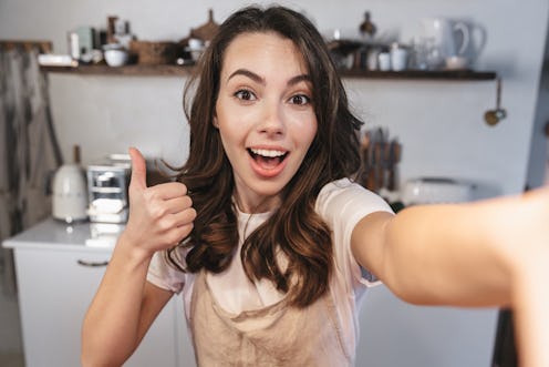 Attractive brunette woman wearing apron taking a selfie while sitting at the kitchen at home, thumbs...