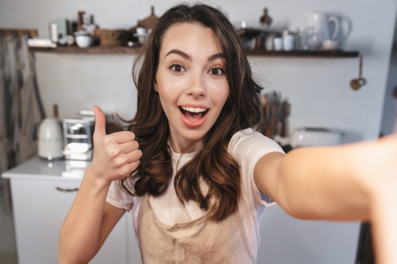 Attractive brunette woman wearing apron taking a selfie while sitting at the kitchen at home, thumbs...