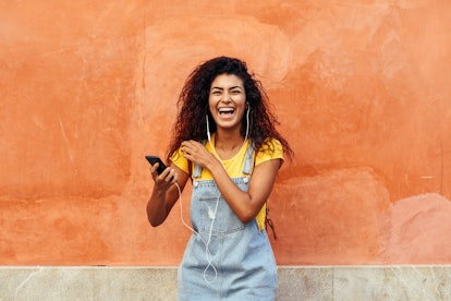 A girl in overalls and a yellow T-shirt laughs while holding her phone outside in front of an orange...