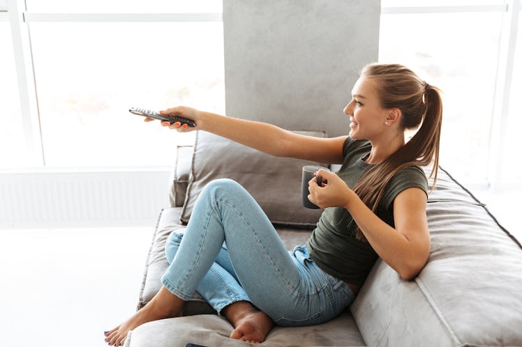 Cheerful young woman sitting on a couch at home, changing channels, drinking tea from a cup