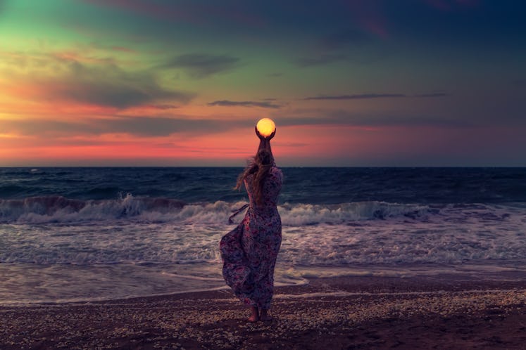 Young woman standing on the beach during sunset, thinking about the April 2022 full Pink Moon on Apr...