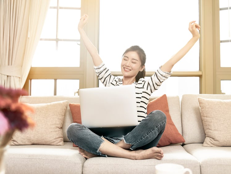 beautiful and happy young asian woman working from home sittng on couch using laptop computer, arms ...