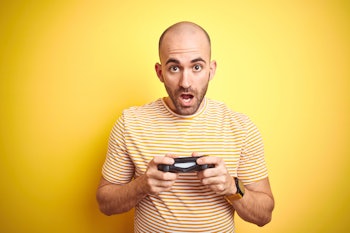 Young man playing video games using joystick gamepad over isolated yellow background scared in shock...