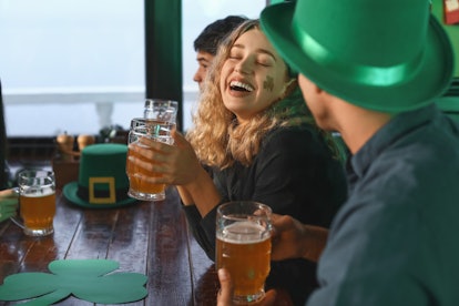 A woman with a shamrock sticker on her cheek laughs while celebrating St. Patrick's Day with friends...