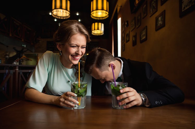 A man and woman hold mojito glasses and laugh at a bar.