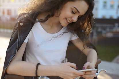 Cropped outdoor shot of pretty girl with charming shy smile reading romantic text message from her b...