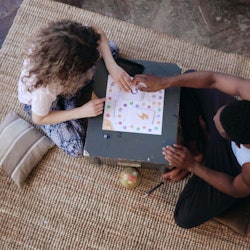Top view of multiracial couple sitting on floor and playing board game. Woman throws dice, takes car...