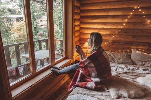 Woman in warm blanket relaxing and drinking morning coffee on cozy bed in log cabin in winter