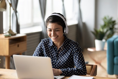 A happy woman wearing headphones and a blue polka dot button down shirt smiles while typing on her l...