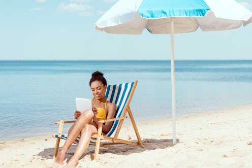 african american woman relaxing on deck chair and using digital tablet under beach umbrella in front...