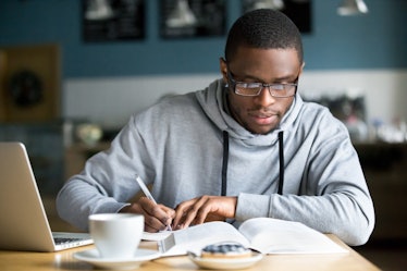 Focused millennial african american student in glasses making notes writing down information from bo...