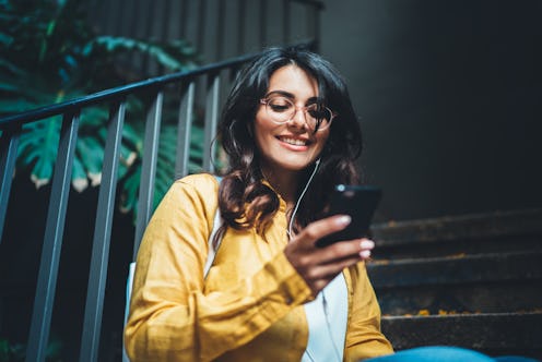 A trendy brunette woman smiles while sitting on the steps of her apartment and texting.