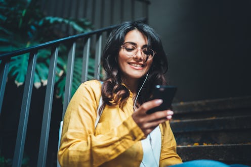 A trendy brunette woman smiles while sitting on the steps of her apartment and texting.