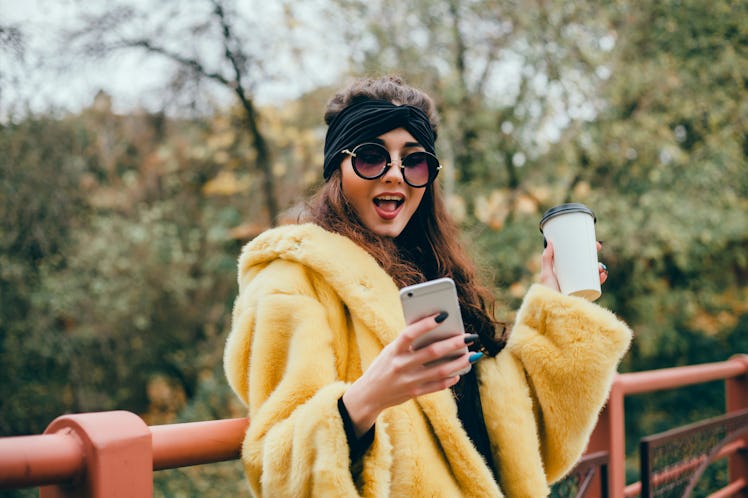 A woman in a yellow faux fur coat, black sunglasses, and a black headband looks at her phone and hol...
